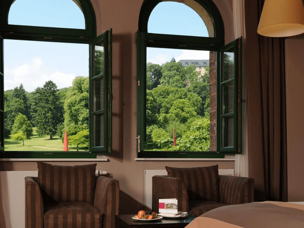 Blick auf zwei geöffnete Fenster mit Aussciht ins Grüne, davor zwei Sessel mit Beistelltisch
