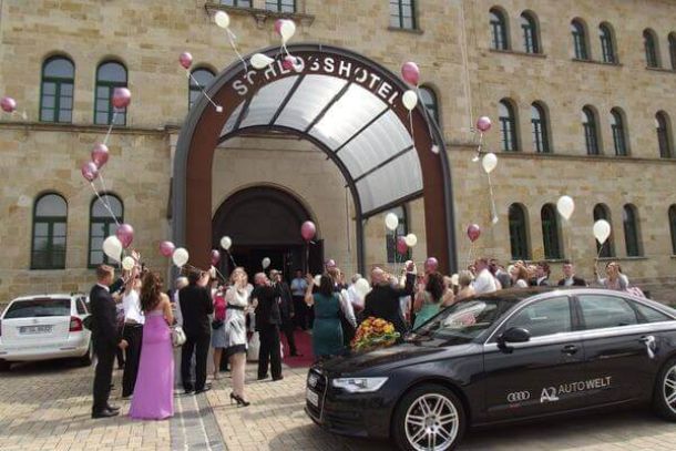 Hochzeitsgesellschaft mit Luftballons und Hochzeitsauto vor dem Hoteleingang