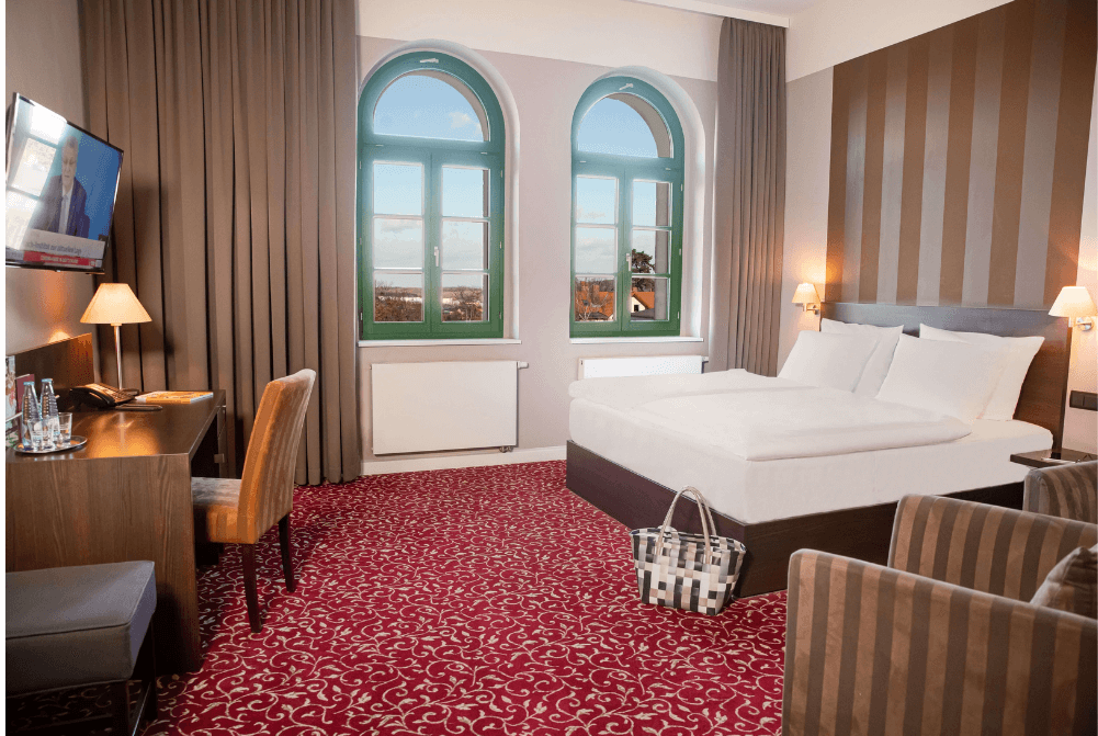 Zimmeransicht mit Schreibtisch, Sessel, TV, Bett, Wellnesstasche und großen Fenstern