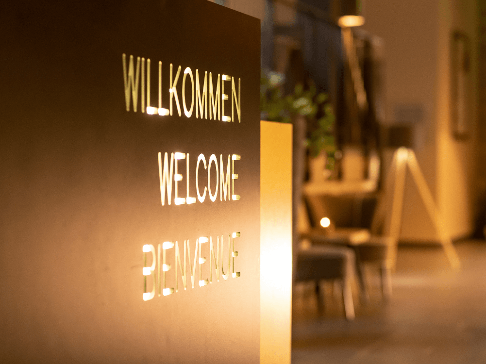 Willkommensschild am Empfang in der Hotellobby mit dem Wort Willkommen in Deutsch, Englisch und Französisch