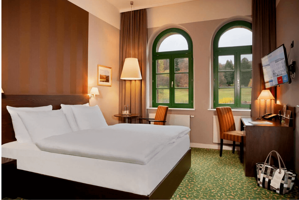 Zimmeransicht mit großem Doppelbett, Schreibtisch, Stuhl, TV, Wellnesstasche, großen Fenstern und Blick ins Grüne