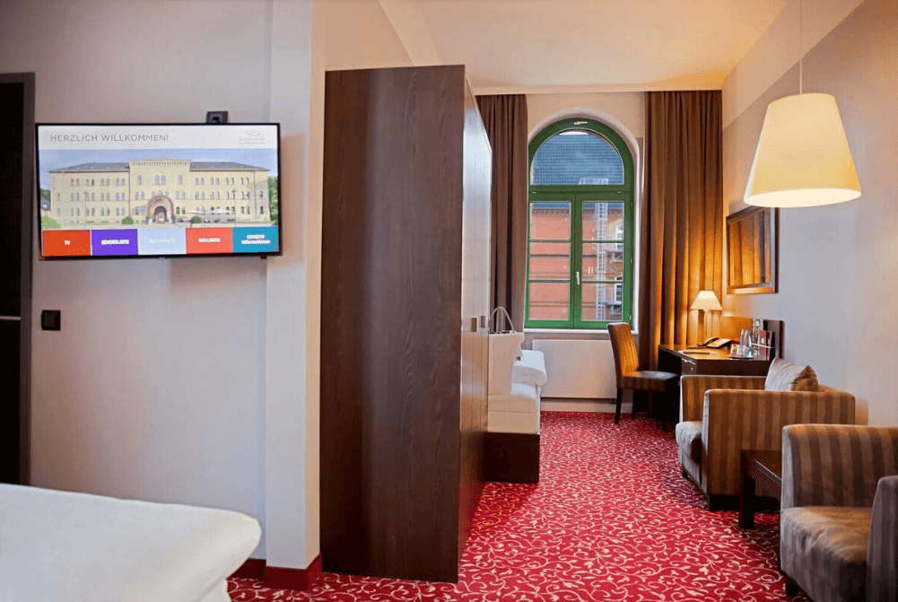 Zimmeransicht mit Doppelbett, Nische mit Einzelbett, Schreibtisch, zwei Sessel, großem Fenster und TV