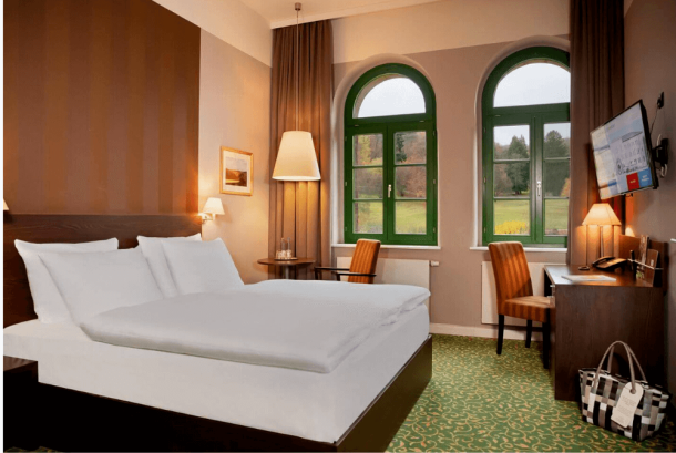 Zimmeransicht mit großem Doppelbett, Schreibtisch, Stuhl, TV, Wellnesstasche, großen Fenstern und Blick ins Grüne