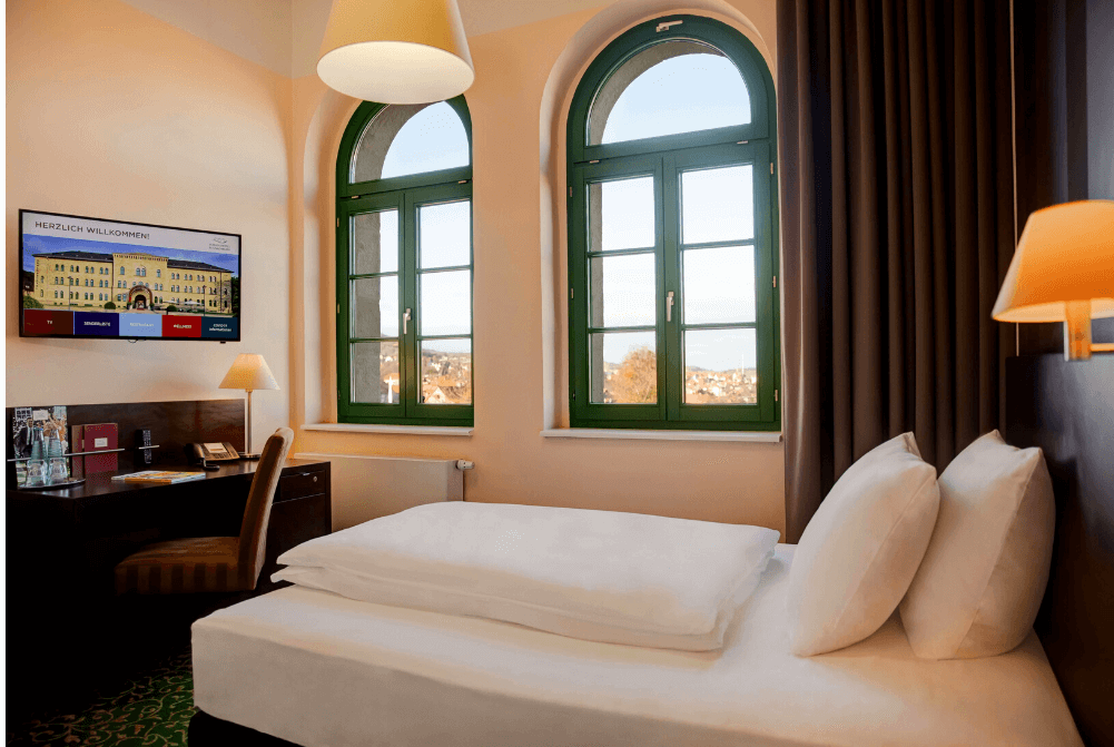 Zimmeransicht mit einem schmalen Bett, Schreibtisch, Stuhl, TV und großen Fenstern