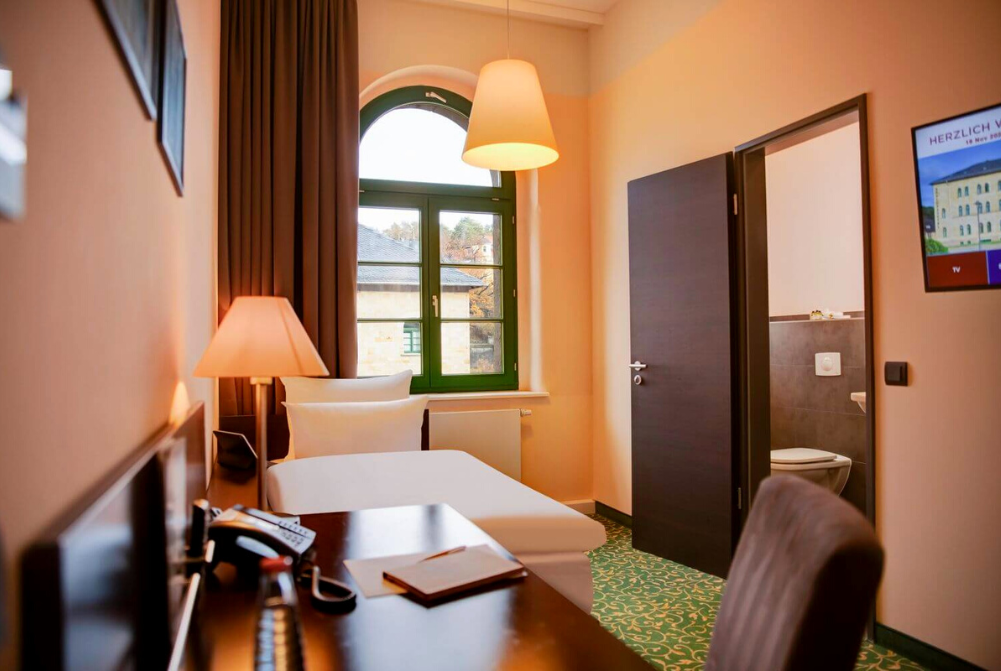 Zimmeransicht mit einem Einzelbett, Schreibtisch, Stuhl, großem Fenster und Blick ins Badezimmer